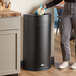 A woman putting a bag of food in a Rubbermaid black steel half round trash can.