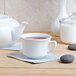 A white Tuxton China cup of tea and a white teapot on a table.