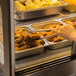 A person using tongs to serve food from a Vollrath countertop heated display case.