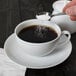 A person using a spoon to pour sugar into a white 10 Strawberry Street porcelain cup.