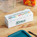 A white Noble Products dispenser with 7 different colored date labels on a wooden surface.