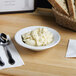 A Tuxton bright white china bowl filled with mashed potatoes on a table with a spoon and a basket of bread.