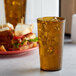 An amber plastic tumbler filled with a brown liquid and ice on a table with sandwiches.