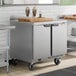 A stainless steel two-door worktop freezer on wheels with a cutting board and milkshakes on top.