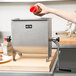 A person using a Weston Manual Meat Mixer to pour sauce from a bottle into a plastic container.
