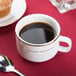 A CAC bright white porcelain coffee cup filled with coffee on a red table surface with a spoon.