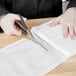 A person cutting a piece of white San Jamar Grade 40 bleached cheesecloth with scissors.
