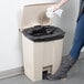 A person's leg in jeans and boots standing next to a Rubbermaid beige rectangular step-on trash can with a black bag inside.