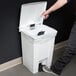 A person's hand reaching to open a white Rubbermaid step-on trash can.