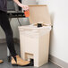 A woman using a Rubbermaid beige rectangular step-on trash can to throw away a coffee cup.