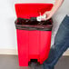 A person throwing a paper into a Rubbermaid red rectangular step-on trash can.