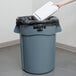 A hand throwing a white container into a grey Rubbermaid commercial trash can.