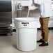 A man in a chef's uniform pouring flour into a large white Rubbermaid trash can.