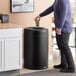 A man in a purple shirt throwing a cup into a Rubbermaid Crowne round open top trash can.