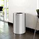 A silver Rubbermaid Atrium waste receptacle in a room.