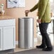 A person in a yellow shirt throwing a cup into a Rubbermaid Atrium satin finish trash can.