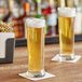 Two Acopa Pilsner glasses of beer on a table.