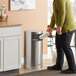 A man standing next to a Rubbermaid metallic round satin stainless steel trash can.