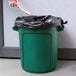 A hand putting a black plastic bag into a green Rubbermaid commercial trash can.