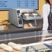 A woman in a striped apron standing in front of a Hatco Intelligent Toast-Qwik dual conveyor toaster in a bakery.