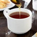 A spoon pouring liquid into a Tuxton Modena AlumaTux Pearl White cup of tea.