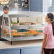 A woman in a pink shirt standing in front of a Hatco stainless steel countertop hot food merchandiser.