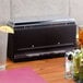 A black rectangular Vollrath Straw Boss dispenser on a counter with a glass of water next to it.