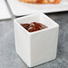 A white square Tuxton ramekin with brown sauce in it.