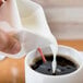 A person pouring milk into a white Tuxton AlumaTux creamer.