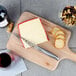 A Franmara stainless steel serrated cheese knife cutting cheese on a cutting board next to crackers and a bottle of wine.