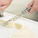 A person using a Franmara stainless steel serrated cheese knife to cut cheese on a white cutting board.