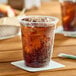 A clear 12 oz plastic cold cup filled with a dark carbonated beverage and ice, placed on a coaster.