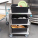 A Lakeside stainless steel utility cart with black containers holding white and green plates.