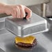 A person using an American Metalcraft square aluminum basting cover to press a cheeseburger on a grill.