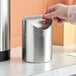 A hand putting a red straw into an American Metalcraft stainless steel swing waste bin on a counter.