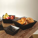 A black square bread basket filled with brown and white bread on a table with a bowl of fruit.