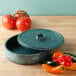 A round green polyethylene container with a lid on a table filled with tomatoes.