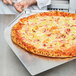 A hand holding a silver object cuts a pineapple pizza on a metal tray.