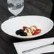 A white 10 Strawberry Street Whittier shallow oval porcelain bowl on a table with a glass of water and a long silver spoon.
