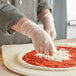 A person wearing Noble Products small powder-free disposable clear vinyl gloves making a pizza.