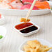 A person using chopsticks to dip vegetables into white sauce in a 10 Strawberry Street divided sauce dish.