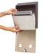 A hand holding a Rubbermaid black steel rectangular cigarette receptacle with a metal mesh.