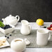 A white pitcher on a table with white dishes and fruit.