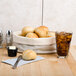 A table with a basket of bread, rolls, and a knife next to a glass of ice tea.