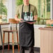 A man wearing a black Chef Revival bib apron holding a tray with bottles and glasses.