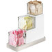 A white metal three tier Cal-Mil glass jar display with stainless steel accents holding condiments.