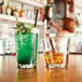 A glass of whiskey with ice next to a clear Prysm glass filled with a green and brown drink.