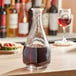 An Arcoroc square glass carafe filled with dark liquid on a table with a glass of red wine.