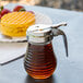 A Tablecraft beehive glass syrup dispenser with brown syrup in it sits on a table next to waffles.