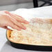 A hand using a plastic container to put food in a black and gold aluminum foil pan with a dome lid.